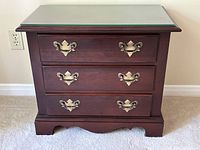 Front view of the cherry wood end table showing three drawers with brass handles and glass top, placed against a wall over carpet.
