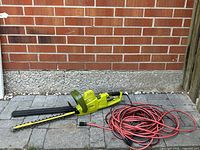 Sun Joe electric hedge trimmer with blade cover and long extension cord laid on stone pavement against brick wall.