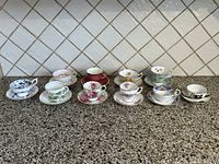Front view of the 10 assorted fine china teacups and saucers arranged in a single line on a granite countertop.