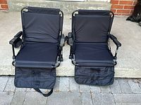 View of two black foldable lawn chairs side by side with footrest flaps unfolded, showing padded backrests, seats, and armrests with cup holders.
