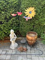 Photo of entire lot outside on stone patio against a green hedge, showing colorful metal garden stakes, clay planter on stand, white plaster statue, cast metal decorative items, and hanging hardware