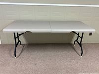 Front view of beige folding card table fully open, showing plastic top and folded metal legs.