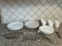 Full set of assorted white plates, mugs, and Lagostina cutlery arranged on countertop against tiled backsplash.