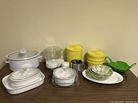 Rival crockpot, food steamer, Corningware bakeware with lids, plastic storage containers, mixing bowls, colander, metal cup and plates on table