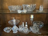 Various crystal serving dishes and bowls displayed on glass shelves including decanter, creamers, salt and pepper shakers, candle holder and more.