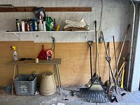 Wide view of garage wall showing garden tools leaned against wooden panel and table with sprays and accessories on top, including garden sprays, hose, and small tools.