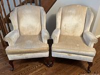 Pair of matching wingback armchairs with beige upholstery and dark wooden legs, shown side by side on wood floor