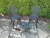 Two black metal garden chairs and one matching metal stool/table placed on stone patio in front of greenery.