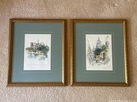Pair of two original watercolour paintings side-by-side, showing Parisian scenes in gold-tone frames with teal mats laid on a carpeted floor.