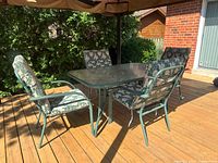 Patio dining set shown on wooden deck with rectangular glass top table and four cushioned chairs in floral pattern. Natural outdoor lighting reveals pale green/blue metal frame.