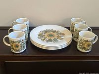 Image of 6 mugs and 4 plates with floral sunflower design on white ceramic, arranged on wooden surface.