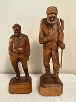 Two wood carved bearded men figurines standing side by side on a white textured surface, showing full front view.