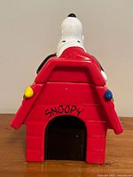 Front view of red ceramic doghouse tea canister with Snoopy lying on top and SNOOPY text on door.
