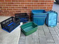Wide shot showing all storage bins and lids together on pavement outside a brick wall, including blue large bins and stacked lids, green bin, and black crates.