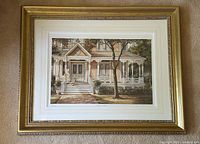 Full view of the print featuring a Victorian house with a porch, framed in gold with white matting, laid flat on carpet.