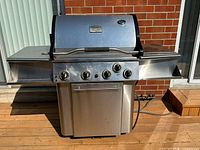 Frontal image of the Vermont Castings Signature Series barbecue grill on a wooden deck, showing stainless steel construction, four control knobs, attached side shelves, and temperature gauge on the lid.