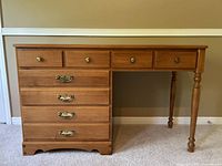 Full front view of the vintage maple desk showing four small top drawers with brass knobs and four larger drawers with ornate brass pulls, turned leg on right, scalloped base trim.