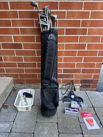 Full frontal view of the vintage right-handed golf lot including black golf bag with clubs against red brick wall, and assortment of golf accessories laid out on the ground.