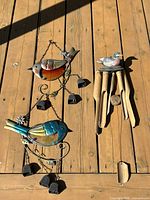 Photo showing two metal and colored glass bird wind chimes each with hanging metal bells, and one carved painted wooden bird wind chime with wooden hanging tubes.