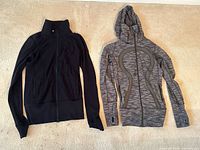 Photo showing two women's Lululemon Define jackets laid flat on the floor: a solid black funnel neck jacket with zip front and a salt and pepper grey hooded jacket with zip front and thumb holes.