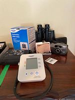 Blood pressure monitor device, cuff, and original box with black binoculars and Pentax camera with case on wooden surface