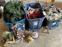 Three blue plastic storage bins filled with artificial greenery, red berries, white poinsettias and a large coil of white string lights. Several Christmas decorations including a snowman sled, small planter box, and various ornaments are placed in front of the bins.