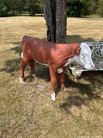 Concrete cow statue shown outdoors in a grassy area next to a weathered white metal bench and a tree, facing sideways showing full body.