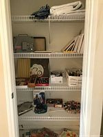 Photo of open closet showing shelves with various art and craft supplies including canvases, boxes with paints and brushes, folded fabric, and other materials.