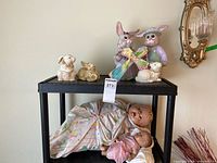 Full lot view showing large worn composite doll in floral pajamas with small composite doll below. Easter figurines on top of shelf: various rabbits and chick.