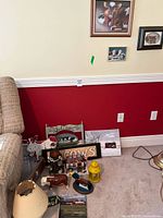 Photo showing assortment of folk art decor items including painted signs, wooden animal figurines, small lantern, and framed cow-themed artwork on floor against wall with red lower half and beige upper half.