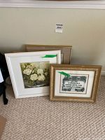 Lot of framed artworks leaning against a wall on carpet, includes floral painting, embroidered quote, and another piece with gold fabric-textured mat frame.