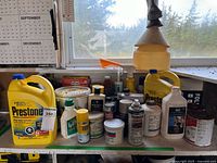 Photo showing a variety of garage liquids including two large Prestone antifreeze jugs, various small bottles, cans and a white funnel on a workbench under a window