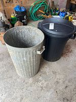 Two garbage cans side by side: one metal galvanized can and one black plastic can with lid.