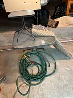 Two metal garden tables stacked, with a rolled outdoor mat and coiled garden hose with nozzle on the floor.