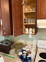 Photo of kitchen cabinet showing glassware and coffee mugs on shelf and countertop, includes clear glass serving bowls and floral mugs