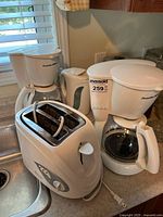 View of two Proctor Silex coffee makers, Betty Crocker toaster, and electric kettle on countertop near kitchen sink.