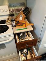 View showing wooden knife block with black-handled knives, cutting board with fruit design on countertop, stove and refrigerator visible, drawer open with assorted silverware and cooking utensils.