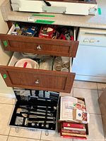 Two drawers open showing kitchen utensils, cookbooks, glass casserole dishes, wooden utensils, and a black BBQ toolset in a black carrying case.