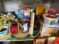 Photo showing inside of cabinet containing various electrical and hardware items such as wires, rope, containers with nails, power strip, and light bulb boxes.