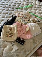 Overview of assorted textile items including baby clothing, slips, handkerchiefs, opera glasses on mattress