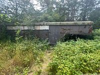 Side view of livestock trailer showing extensive rust and wear with a mostly flat roof and surrounding greenery.