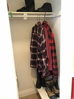 Closet view showing three men's flannel shirts hanging, including red and black plaid, dark blue and white plaid, and a solid color shirt, plus four pairs of men's boots on floor beneath.