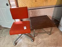 Office chair with orange upholstered seat and back and metal wheeled base next to wooden side table.