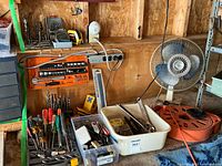 Wide shot showing assortment of hand tools, drill bits, extension cord reel, oscillating fan, and more laid out on a workbench in a workshop setting.
