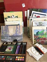 Stacks of assorted canvases with various artwork, several partially finished, leaning against a wall. A wooden art supply case containing colored pencils, pastels, watercolor sets, and brushes along with white mixing palettes is on the floor in front of canvases.