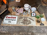 Wide view of assorted vintage and antique household and collectible items arranged on plywood floor showing signs, kitchenware, glassware, and small collectibles.