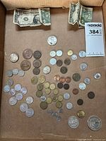 Top view of mixed world coins and paper currency spread on a flat surface, showing the range of coin types and two one-dollar bills.