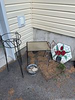 Photo showing the overall lot placed against siding with plant stand, table, basket, bucket with rocks, and mosaic tile