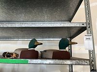 Two handmade wooden duck decoys placed side by side on a metal shelf, showing painted green heads, grey bodies, and brown chests.