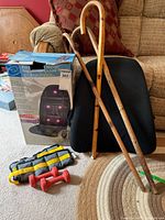 Photo showing black seat massager pad with attached remote, two wooden walking canes, yellow and black ankle weights, and one red hand weight on carpet next to box for massager.
